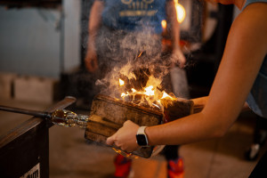 A person with light skin and dark hair works with a piece of molten glass attached to a metal rod in front of a glowing furnace.