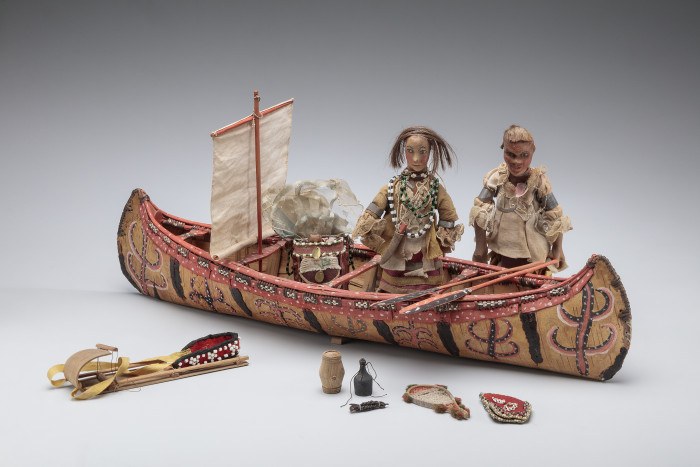 Dolls standing next to a model canoe