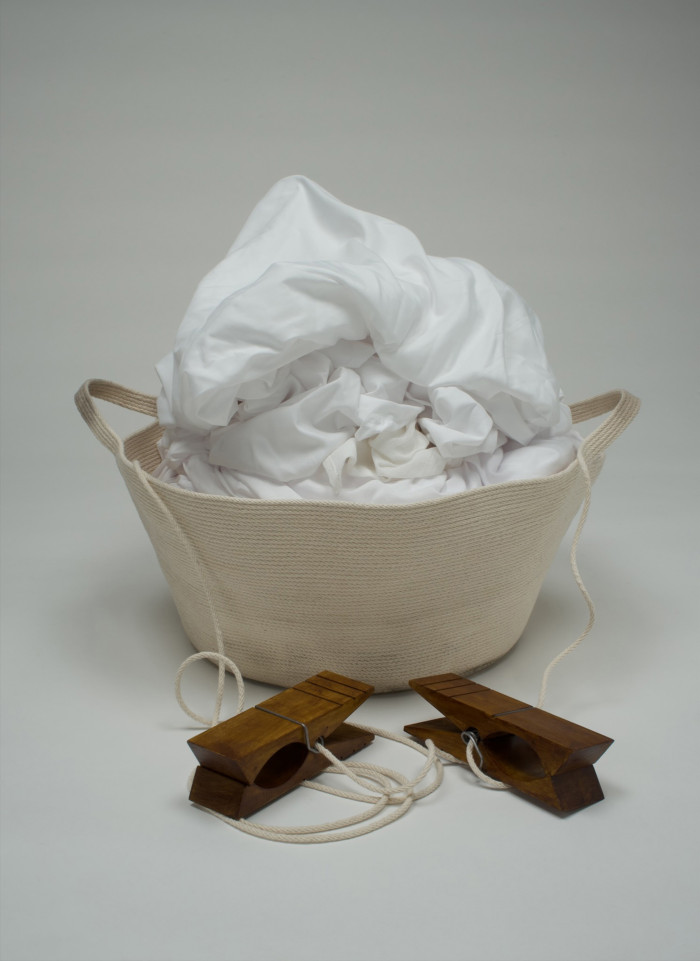 A fabric basket full of white cloth. In front of it sit two oversized dark wood clothespins.